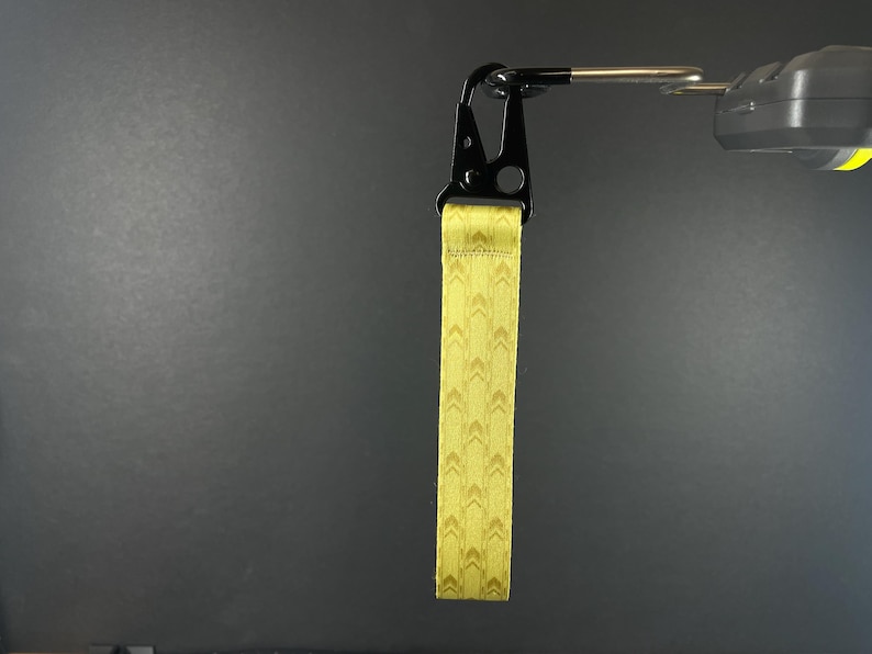 K&ouml;nnte beinhalten: Ein gelbes Stoff-Handgelenkband mit einem sich wiederholenden Chevron-Muster, befestigt an einem schwarzen Metallkarabiner. Das Band besteht aus gewebtem Material mit gen&auml;hten Kanten und h&auml;ngt vertikal an einem silbernen Metallhaken.