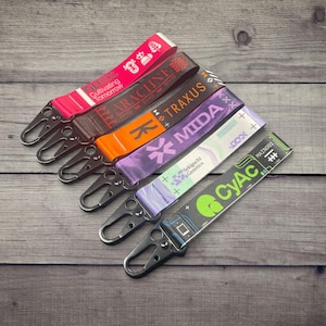 May include: A collection of six colorful lanyards with metal clasps, arranged on a wooden surface. Each lanyard features a unique design with text and graphics in various colors, including red, brown, orange, purple, light blue, and green.