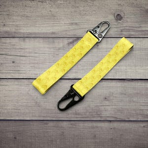 K&ouml;nnte beinhalten: Zwei gelbe Schl&uuml;sselb&auml;nder aus Stoff mit einem sich wiederholenden Chevron-Muster. Jedes Band ist an einem Ende mit einem schwarzen Metallkarabiner ausgestattet. Der Text auf der linken Seite lautet: GREAT FOR: KEYS, BACKPACKS, TOTES, & MORE.