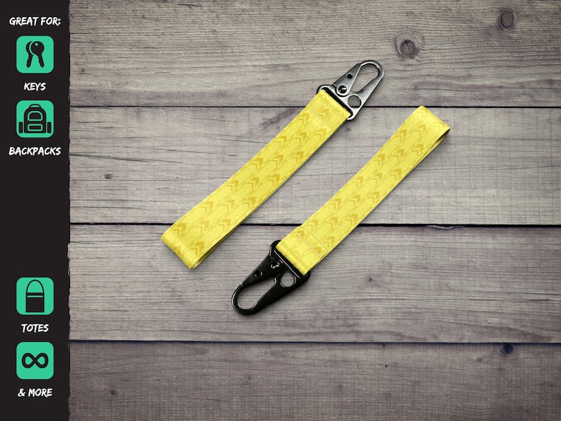 K&ouml;nnte beinhalten: Zwei gelbe Schl&uuml;sselb&auml;nder aus Stoff mit einem sich wiederholenden Chevron-Muster. Jedes Band ist an einem Ende mit einem schwarzen Metallkarabiner ausgestattet. Der Text auf der linken Seite lautet: GREAT FOR: KEYS, BACKPACKS, TOTES, & MORE.