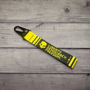 May include: A black and yellow fabric keychain featuring a black metal clip at the top. The strap displays a yellow skull icon and the text LIBERTY. DEMOCRACY. FREEDOM. printed in yellow, finished with yellow stripes and small stars at the end.