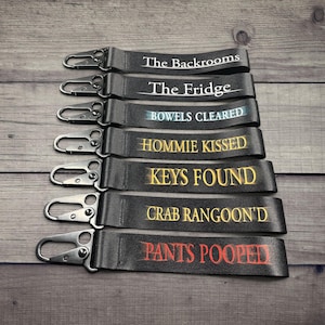 May include: A stack of black keychains with silver clasps, each bearing a different phrase in a unique font. The phrases include "The Backrooms," "The Fridge," and "PANTS POOPED" in red.