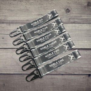 May include: Six gray lanyards with black metal clasps are arranged on a wooden surface. Each lanyard is labeled with "NOBLE" followed by a number and a name and code. A white geometric design is also present.