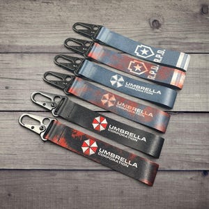 May include: Five keychains with black metal clasps and fabric straps. The straps feature the text "R.P.D." and "Umbrella Corporation" with star and umbrella logos. The straps have a distressed, red and blue color scheme, set against a wooden background.