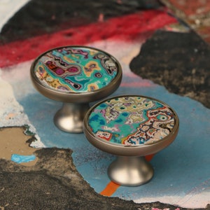May include: Two silver cabinet knobs with a colourful abstract design. The knobs have a round base and a circular top with a swirling pattern of blue, green, yellow, and pink.