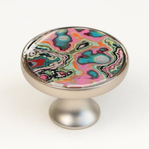 May include: A round, silver cabinet knob with a colorful, abstract design. The design features swirls of pink, blue, green, orange, and black.