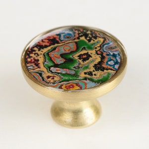 Puede incluir: Un pomo de latón con un diseño redondo, abstracto y colorido. El pomo tiene una parte superior circular y una base redondeada.