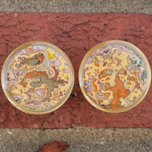 May include: Two circular decorative items with a gold edge. The interior has a colourful, abstract pattern in yellow, orange, pink, and green. They are set on a red brick and grey concrete surface.