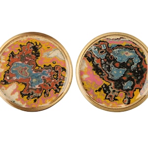 Puede incluir: Dos objetos decorativos redondos con borde dorado y patrones abstractos coloridos en su interior. Los patrones recuerdan a un efecto de mármol en espiral en tonos de rosa, amarillo, azul, negro y rojo.