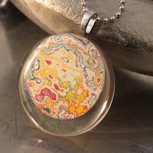 May include: A silver chain necklace with a round pendant featuring a colorful abstract design. The pendant is made of clear resin with a swirling pattern of yellow, red, pink, and blue.