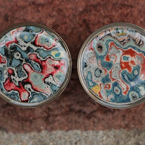 May include: Two circular plugs with silver-coloured rims, showcasing vibrant, abstract patterns. The designs incorporate swirls of red, blue, white, and black, creating a marbled appearance. The plugs are set against a brick backdrop.