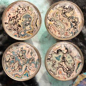 May include: Four round, decorative, gold-colored coasters with a marbled, abstract design in shades of pink, blue, green, black, and white.
