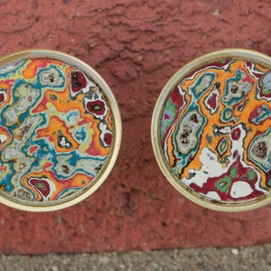 May include: Two round, gold-rimmed coasters with a colorful, abstract, marbled design. The coasters feature a swirling pattern of orange, red, blue, green, and white.