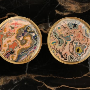 May include: Two round, gold-rimmed, decorative objects with a colourful, abstract, marbled design. The design features swirls of pink, orange, yellow, blue, green, and black.