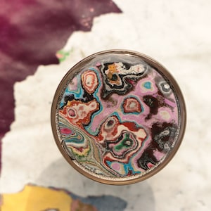 May include: A round, silver-coloured, decorative object with a colourful, abstract design. The design features swirls of pink, blue, green, red, and black.