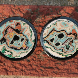 May include: Two round coasters with a black rim and a colourful abstract design. The design features swirls of blue, green, yellow, orange, and black.