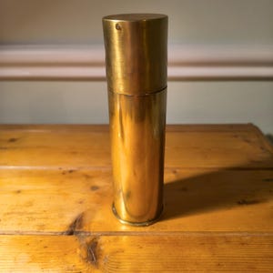 Op de afbeelding: Een cilindrische messing container met een afneembaar deksel. De container heeft een gepolijst, reflecterend oppervlak en staat rechtop op een houten oppervlak. Het messing heeft een warme, gouden tint.