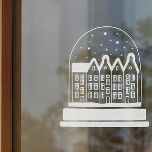 May include: A white silhouette of a snow globe with a row of houses inside. The snow globe is on a window with a wooden frame.