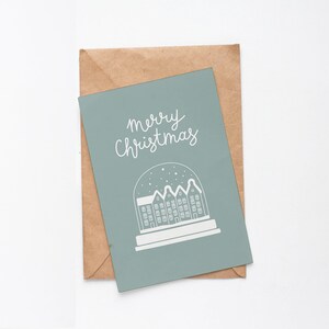 May include: A light blue Christmas card with the text "merry Christmas" in white cursive lettering. The card features a white illustration of a snow globe with a small town inside.