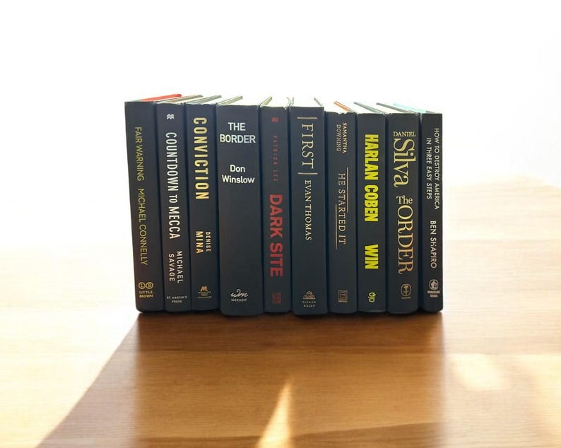 May include: A collection of ten hardcover books with black covers and colorful titles. The books are stacked vertically on a wooden surface. The titles include "Conviction", "The Border", "First", "Harlan Coben", "Silva", and "How to Destroy America in Three Easy Steps".