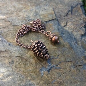 May include: A bronze-colored necklace featuring a detailed pinecone pendant and a small acorn charm. The chain is also bronze, connecting the two nature-inspired charms. The necklace is displayed on a textured, gray stone surface.