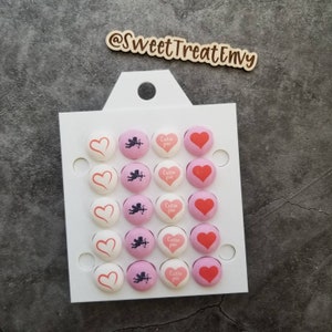 May include: A white card with a cut-out handle holds 20 pink and white chocolate candies. The candies are heart-shaped and feature a Cupid design and the text "Cutie Pie".
