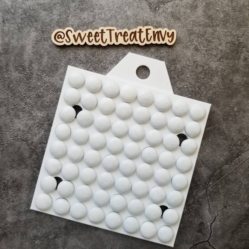 May include: White plastic mold with a handle and a grid of 64 round depressions. The mold is designed for making candy or chocolate. The mold is on a gray textured surface.