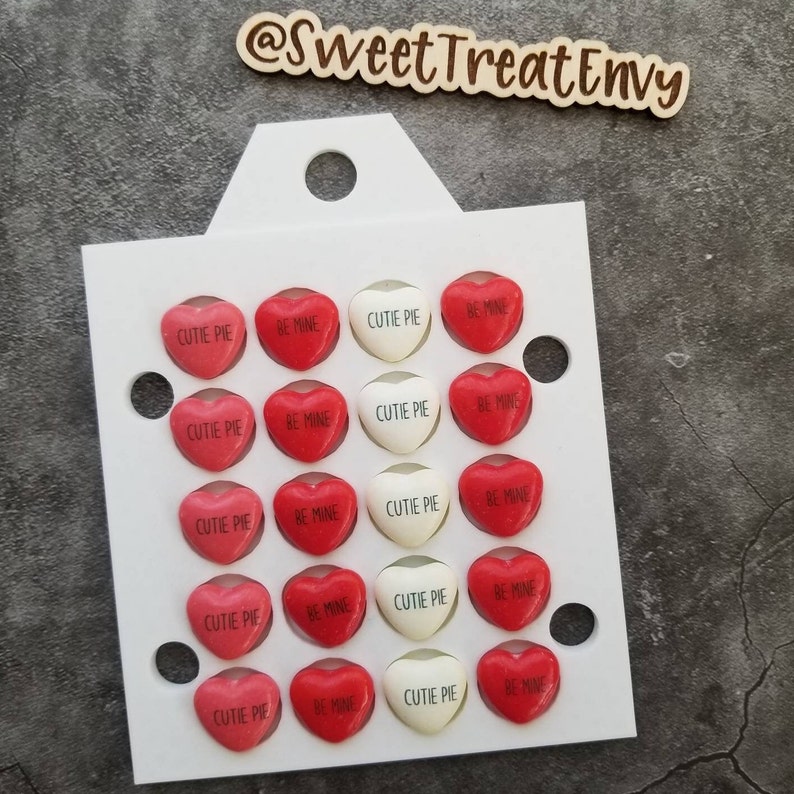May include: A white card with 20 heart-shaped sweets arranged in a grid. The sweets are red and white with the phrases "cutie pie" and "be mine" printed on them.