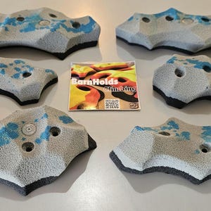 May include: Six gray climbing holds with blue accents and black undersides are arranged on a white surface. A small, colorful card with the text "BarnHolds Climbing" is in the center. The holds have multiple holes for grip.