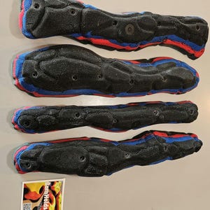 May include: Four black climbing holds with red and blue accents. The holds are textured and have multiple holes for mounting. A small sticker with the text "BarnHolds Climbing" is in the lower left corner.