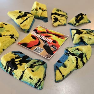 May include: A collection of yellow, blue, and black climbing holds arranged in a circle. The holds have a textured surface and various shapes. A card with the text "BarnHolds Climbing" is in the center.