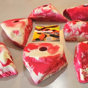 May include: A collection of red and white climbing holds in various shapes, including triangles and rounded forms. Each hold has a hole for mounting and a textured surface. A small card with the text "Barn Holds" is also present.