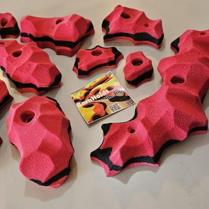 May include: A collection of pink climbing holds with black accents and various shapes. Some holds have holes for mounting. A small card with the text "BarnHolds" is also present.