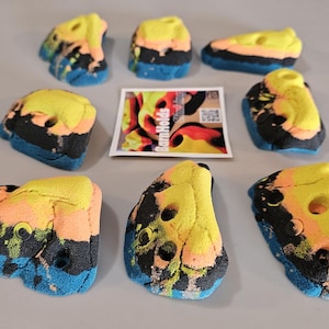 May include: A collection of colorful climbing holds in various shapes. The holds feature a mix of yellow, orange, black, and blue hues. A small card with the text "BarnHolders" is also present.