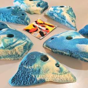 May include: A collection of blue and white climbing holds with a textured surface and a hole for mounting. The holds are in various shapes and sizes, with a marbled appearance. A small card with the text "Climb Holds" is also present.