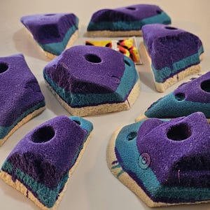 May include: A collection of purple, teal, and beige climbing holds. Each hold has a textured surface, a hole, and a unique shape. The holds are designed for indoor or outdoor rock climbing and bouldering. They are made of a durable material.