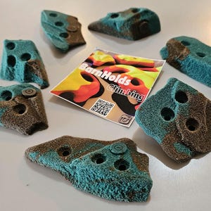 May include: A collection of teal and brown climbing holds with various shapes and sizes. Each hold has multiple drilled holes for mounting. A colorful card with the text "BarnHolds Climbing" is also present.