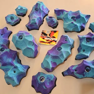 May include: A collection of blue and purple climbing holds with various shapes and sizes. Each hold has a hole for mounting and a textured surface. A sticker with the text "BamHolds" is also present.