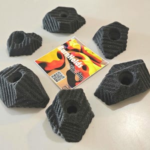 May include: Six black textured climbing holds with a central bolt hole, arranged in a circular pattern around a square sticker featuring the text BarnHolds Climbing and a QR code. Each hold has a ridged surface and an irregular, angular shape.