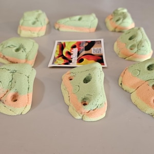 May include: A collection of colorful climbing holds in various shapes. The holds are primarily green with orange and yellow accents. Some have holes for grip. A small card with colorful graphics is also present.