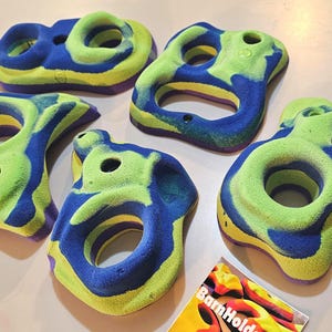 May include: Six colorful climbing holds in various shapes. The holds are a mix of blue, green, and yellow, with a textured surface. Each hold has a hole for mounting. A small card with the text "BarnHolders" is also visible.