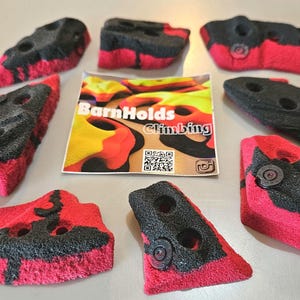 May include: A collection of red and black climbing holds with various shapes and sizes. The holds have a textured surface and are designed for indoor or outdoor climbing walls. A sticker with the text "BarnHolds Climbing" is also present.