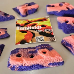 May include: A collection of pink and purple climbing holds with various shapes and sizes. The holds have a textured surface and multiple holes for grip. A sticker with the text "BarnHolds Climbing" is also present.