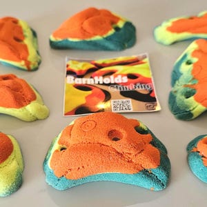 May include: Nine colorful climbing holds in various shapes and sizes, with orange, blue, and yellow hues. A small card with the text "BarnHolds Climbing" is in the center. These holds are designed for rock climbing.