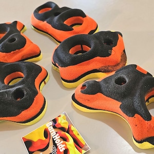 May include: A collection of orange and black climbing holds with yellow accents. The holds are in various shapes and sizes, designed for rock climbing. A small card with the text "BarnHolds" is also present.