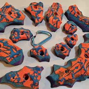 May include: Ten colorful climbing holds with a blue carabiner. The holds are made of a textured material and have a variety of shapes and sizes. They are painted in shades of orange, blue, and purple.