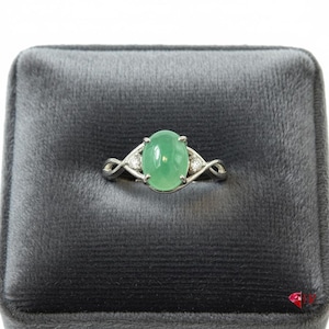 May include: A silver ring with a large oval, light green gemstone as the centerpiece. The ring has a twisted band design with small, clear accent stones on either side of the main stone. The ring is displayed on a gray velvet surface.