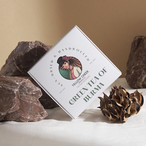 May include: A white box with green and black text that reads "Dried Hand Rolled Sun-Dried Yellow Cheek Green Tea of Burma". The box features an illustration of a woman wearing a traditional Burmese outfit.