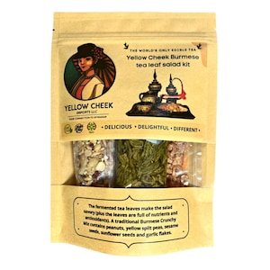 May include: A brown paper bag with a yellow label that reads "Yellow Cheek Burmese tea leaf salad kit". The label also features a picture of a woman in traditional Burmese attire and a tea set. The bag contains three small clear bags filled with various ingredients for a salad.