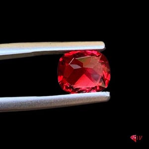 May include: A faceted, cushion-cut, red gemstone is held by tweezers against a black background. The gem has a brilliant, reflective surface, showcasing its internal structure and color. The tweezers are silver.